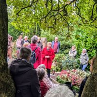 Eine FRau in rotem Mantel steht vor einigen Grabstellen. Sie ist von anderen Personen umringt, die ihr scheinbar zuhören. Im Hintergrund sieht man viele Bäume und Sträucher.