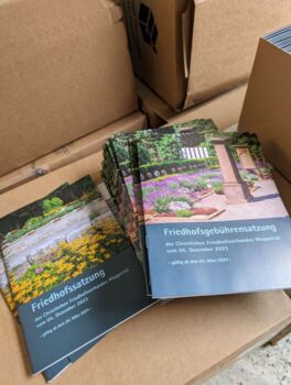 Einige gedruckte Exemplare der Friedhofs- und Gebührensatzung liegen auf braunen Versandkartons.