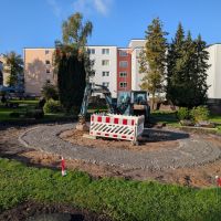 Auf einer Wiese ist kreisförmig eine Vertiefung ausgehoben worden. In dieser Vertiefung steht ein Bagger. Vor dem Bagger wurde ein kleiner Bereich mit rot-weißen Barken abgesperrt.