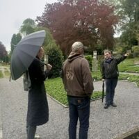Bild einer Führung. Im Vorgergrund sieht man eine Frau und einen Mann von hinten. Diese schauen einen Mann im Hintergrund an, der mit dem Finger auf etws außerhab des Bildes zeigt. Die Personen stehen auf einem Grabfeld.