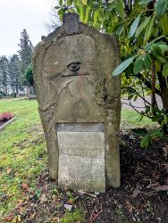 Historische Grabsteinsäule auf einem Friedhof. Oben auf dem Stein ist ein Dreieck mit einem strahlenden Auge, darunter lehnt die in der Mitte zerbrochene Platte mit den Lebensdaten.