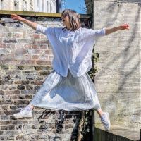 Seitenansicht einer jungen Frau in weißer Bluse und silbernen Rock, die einen großen Schritt von einer kleinen Mauer macht. Sie scheint in der Luft zu schweben. Ihre Arme sind weit ausgebreitet. Im Hintergrund sieht man eine Backsteinmauer.