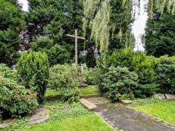 Grabstelle, die mit Rhodedendren und Zypressen bewachsen ist. Auf dem Boden sind Grabsteine eingelassen. Im Hintergrund ist ein hohes Holzkreuz.